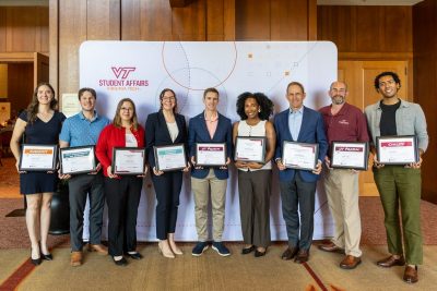 Nine smiling award recipients stand in front of a Virginia Tech Student Affairs-branded backdrop. They are all dressed in professional attire, and each holds their award certificate in front of them.