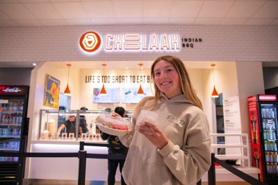 A student holds a to-go container of food and smiles in front of the Choolaah Indian BBQ sign