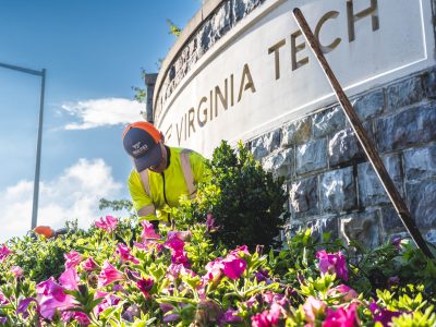 A grounds employee kneels in the dirt while planting a new shrub in front of a gateway wall.