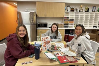Three students smile toward the camera as they pause from writing on cards while sitting around at a table.