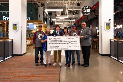 Six smiling people stand holding a large check in a modern dining facility.