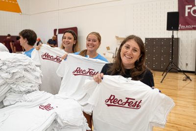 Three people in casual wear standing in front of a table of neatly-stacked white t-shirts. They are smiling and each holding up a white t-shirt that says Rec Fest in red script lettering